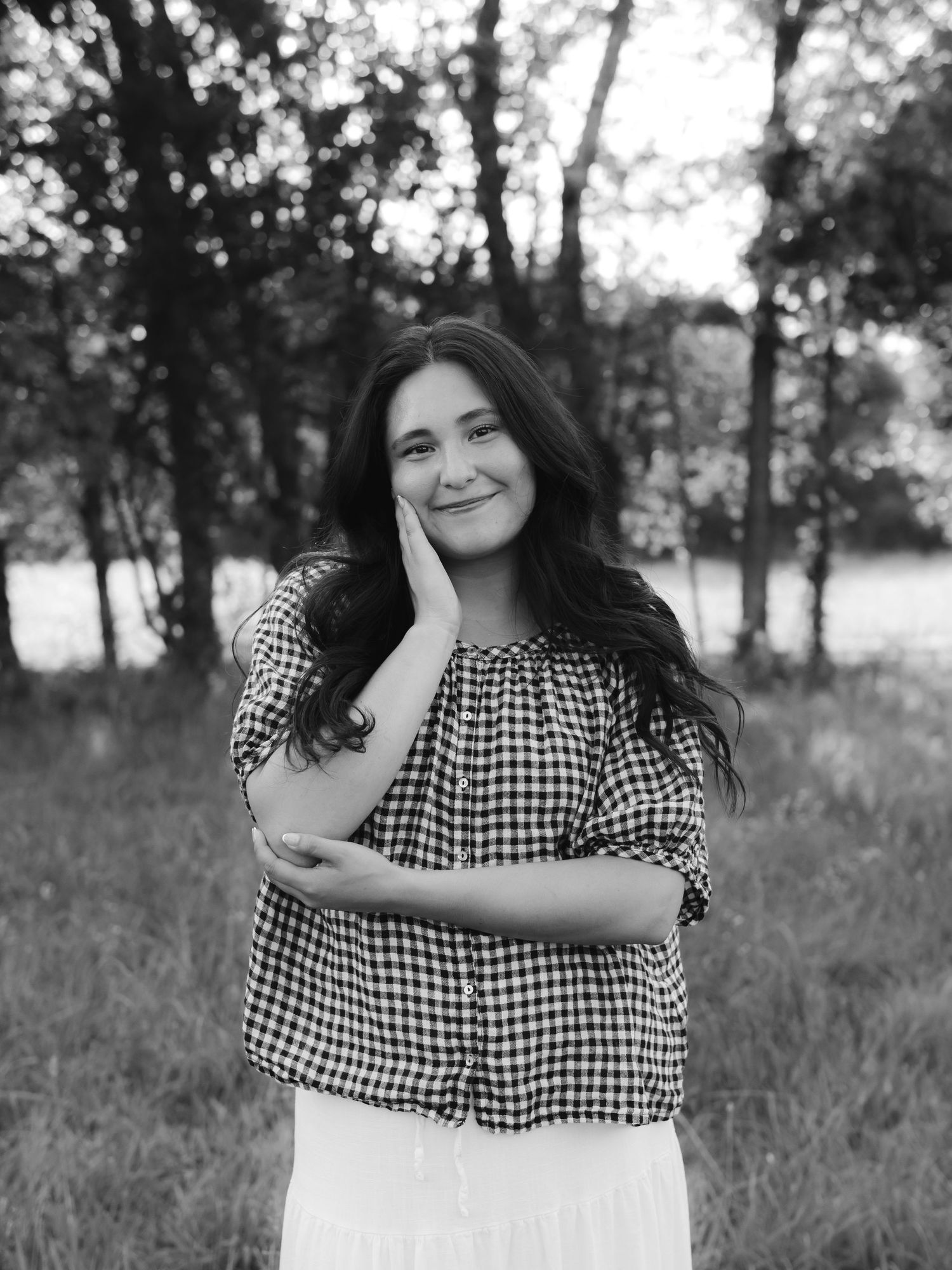 Black and white portrait of a young woman in gingham top smiling outdoors among trees.