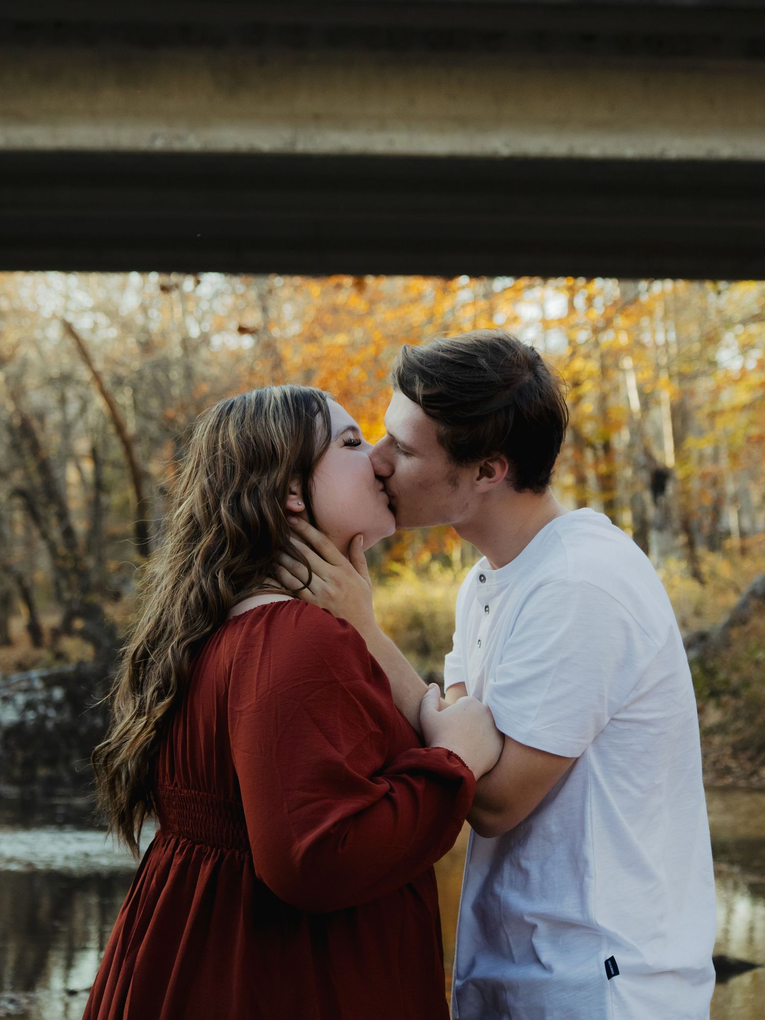A couple shares a romantic kiss under a bridge surrounded by vibrant autumn foliage near a serene river.