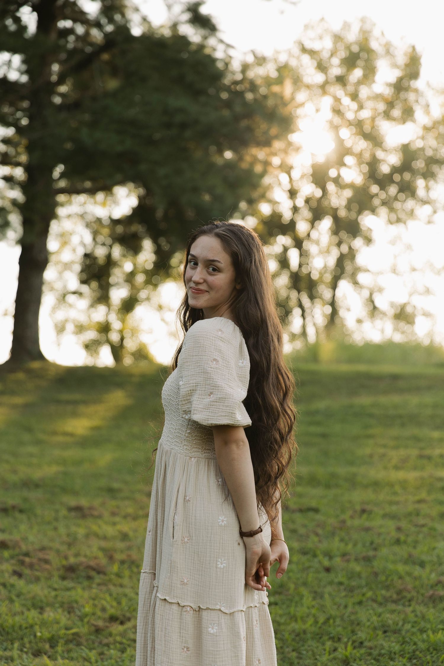 Young woman in white dress standing in sunlit park, long brown hair, smiling over shoulder at golden hour.
