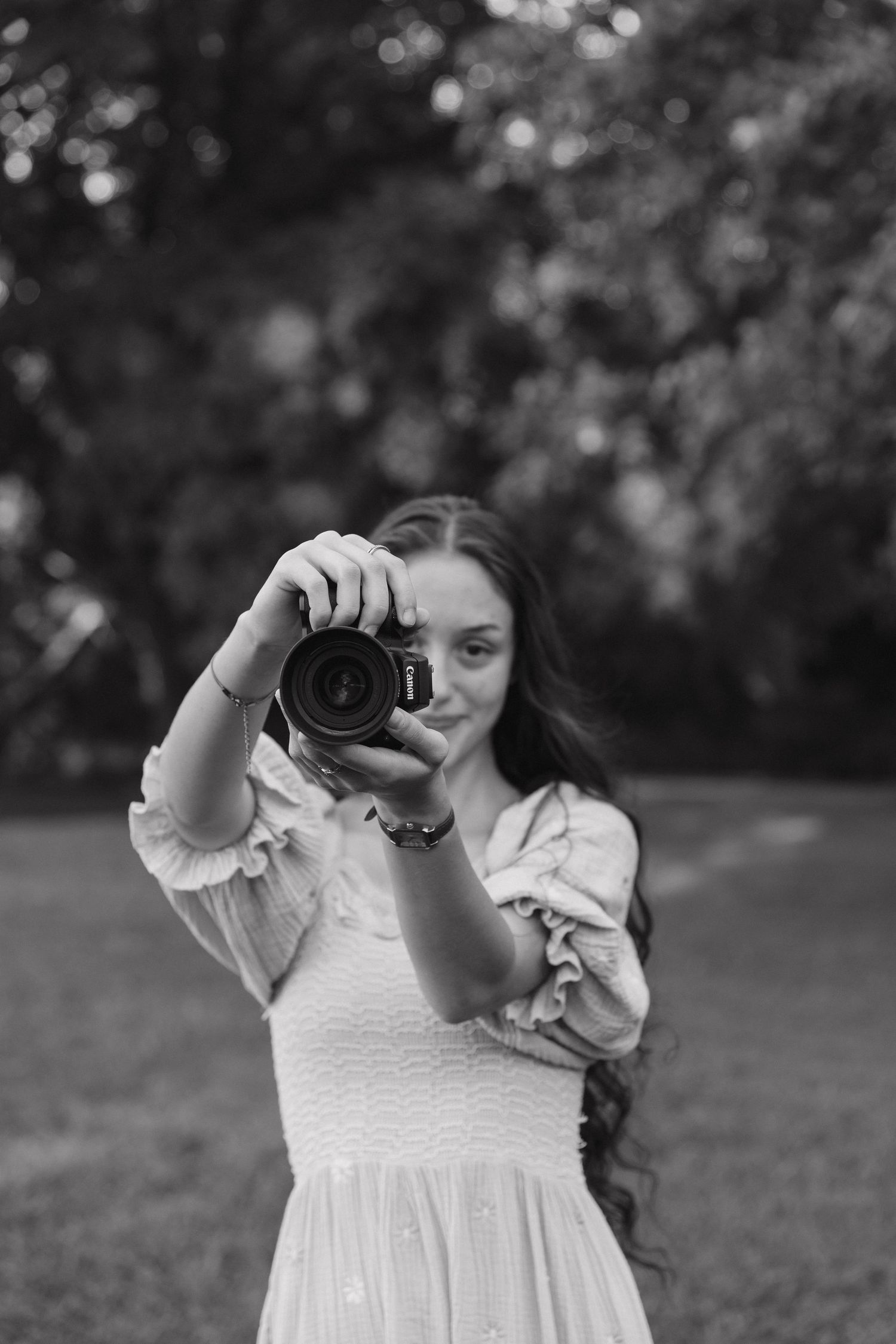 Black and white photo of a young woman in a white dress holding a DSLR camera outdoors in a park setting.