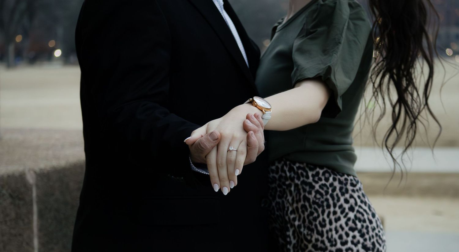 Couple holding hands tenderly, woman wearing leopard print skirt and green top, man in black suit jacket outdoors.