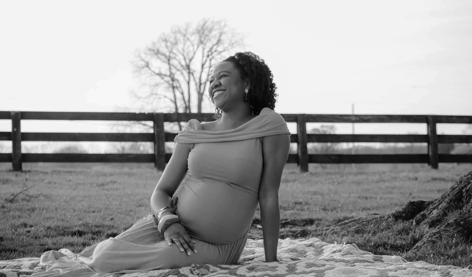 Black and white maternity photo of a pregnant woman sitting gracefully outdoors near a wooden fence.