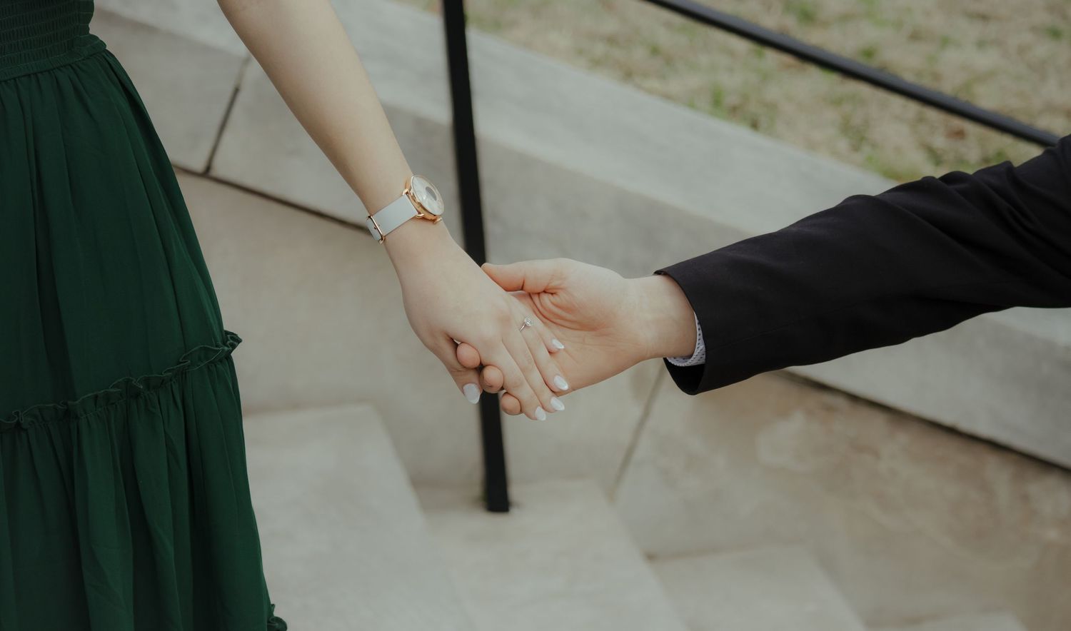 Couple holding hands on stone steps, woman in green dress with watch, man in black suit jacket.