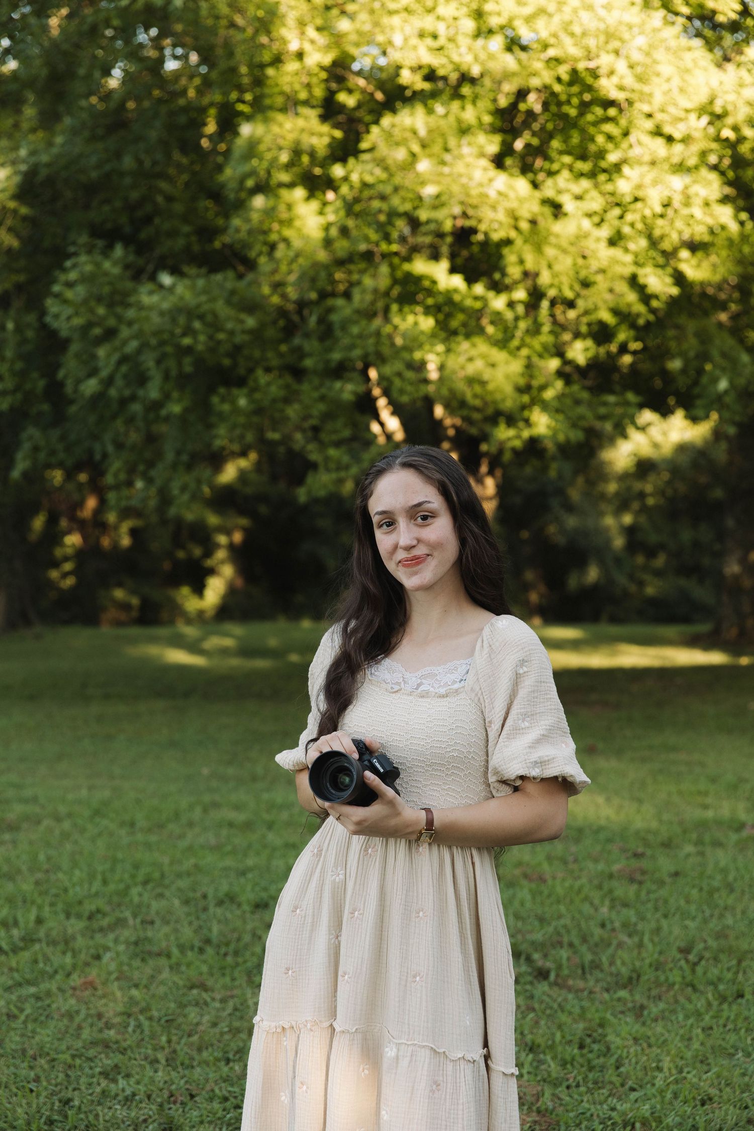 Young woman in cream vintage dress holding a camera, standing on green grass with sunlit trees in background.