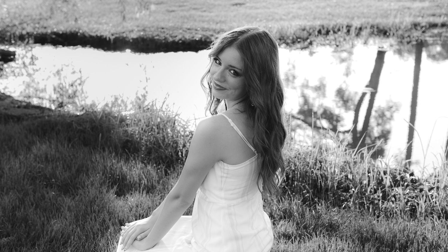 Black and white photo of a young woman in a white dress sitting gracefully on grass near a pond.