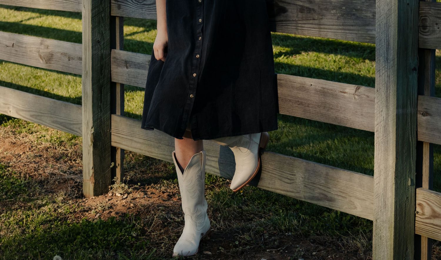 Woman in black dress and white cowboy boots leaning against a wooden fence in a rustic outdoor setting.