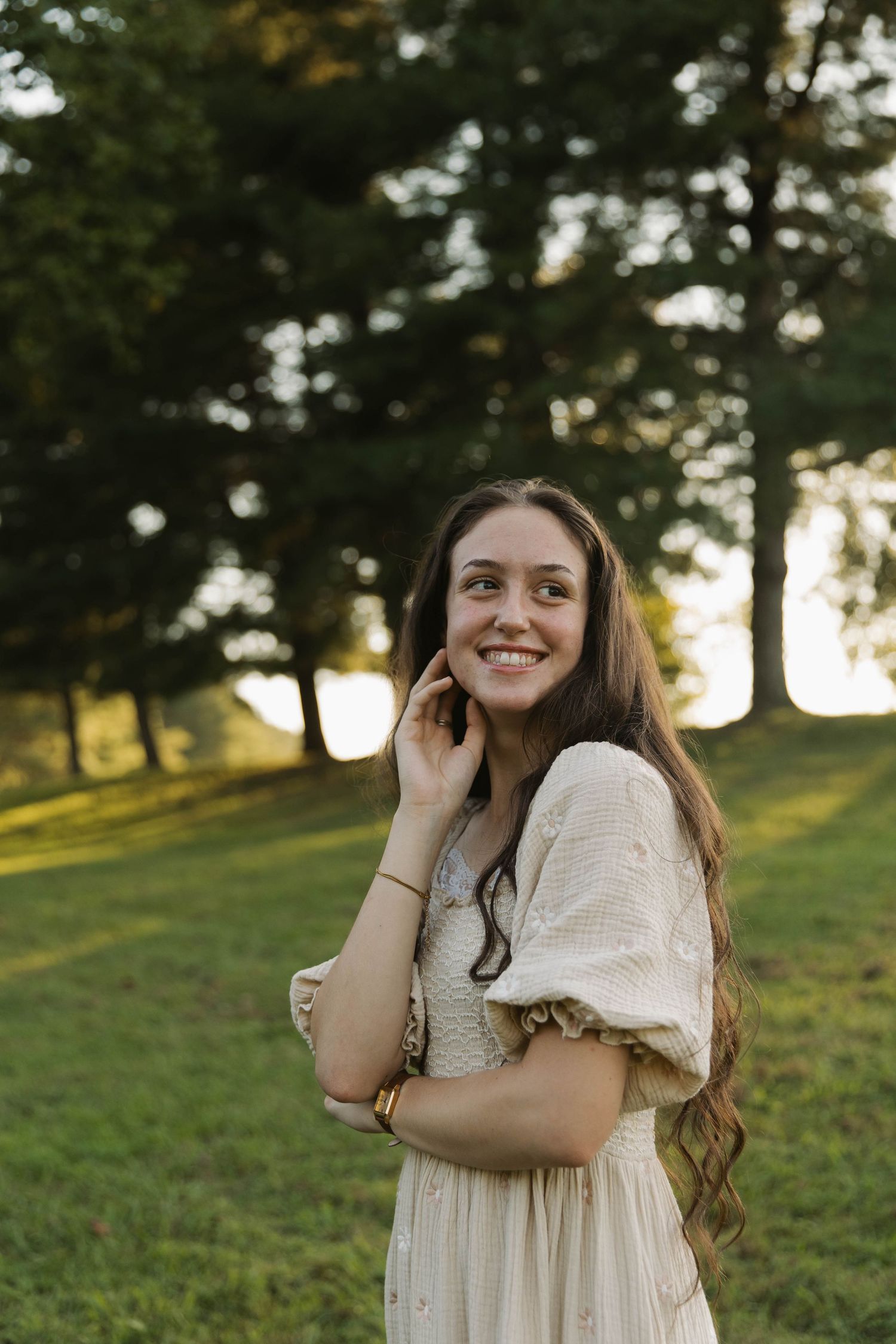 Young woman with long brown hair smiling in a white dress, standing on green grass with trees in the background.