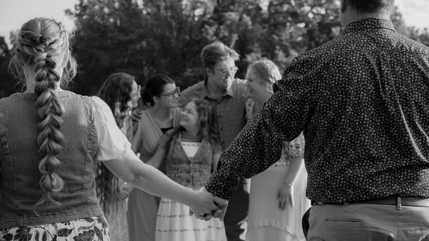 Black and white photo of a group of people holding hands in a circle outdoors at a casual gathering.