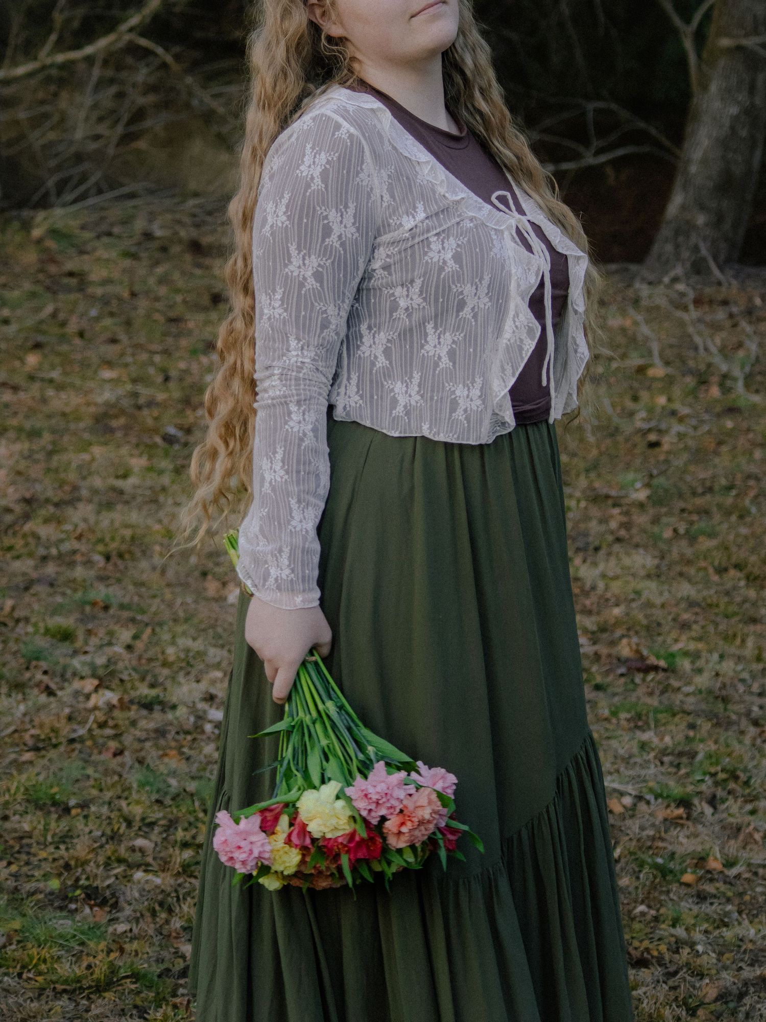 Woman in lace cardigan and green maxi skirt holding colorful bouquet of flowers outdoors.