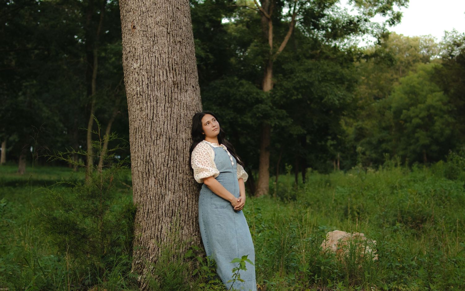 Pregnant woman in blue overalls leaning against a tree in a lush green forest, cradling her baby bump.