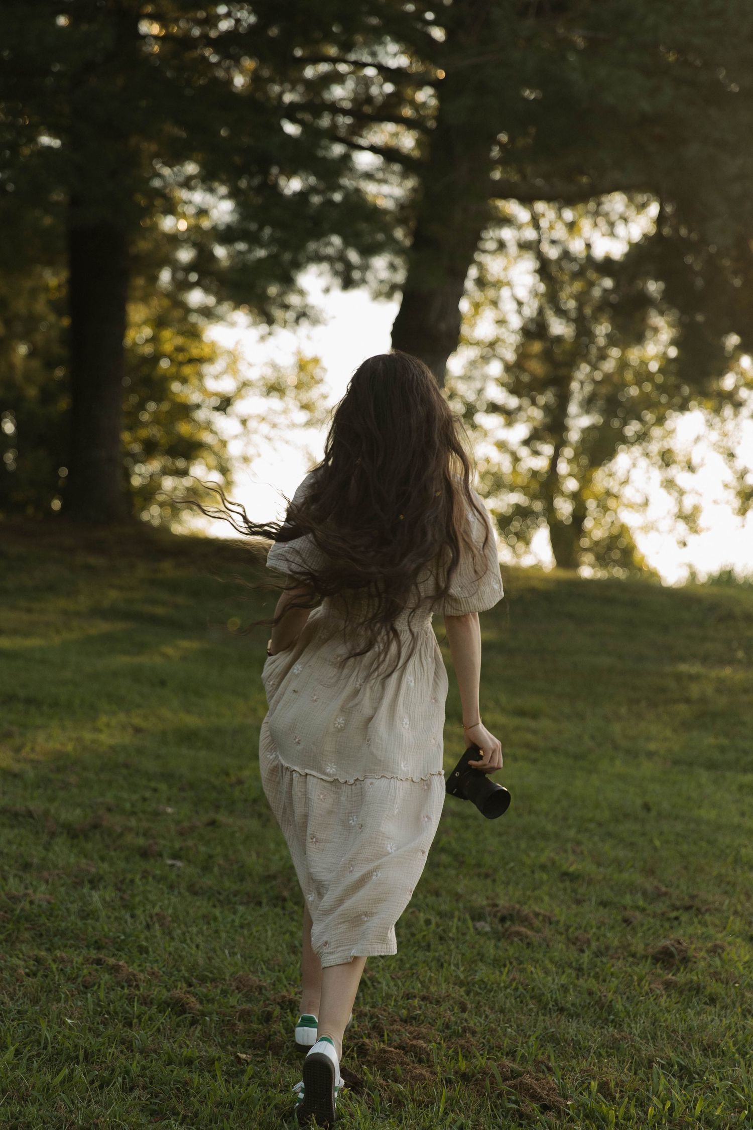 A girl with long brown hair in a white dress runs barefoot across green grass toward sunlit trees.