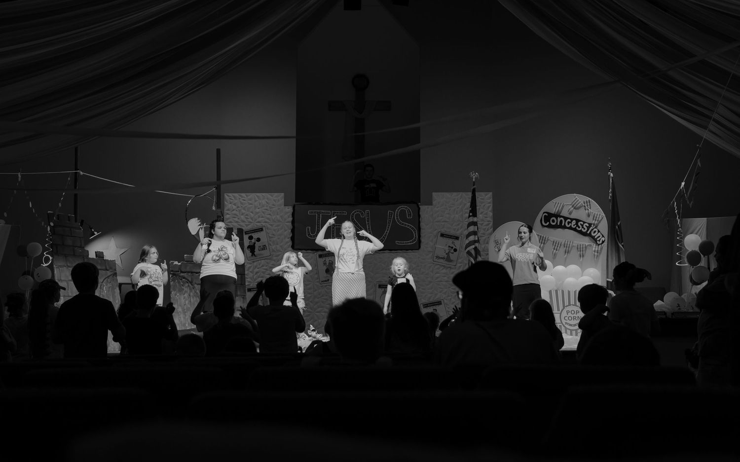 Black and white photo of performers on a lit stage before a silhouetted audience at an indoor event.