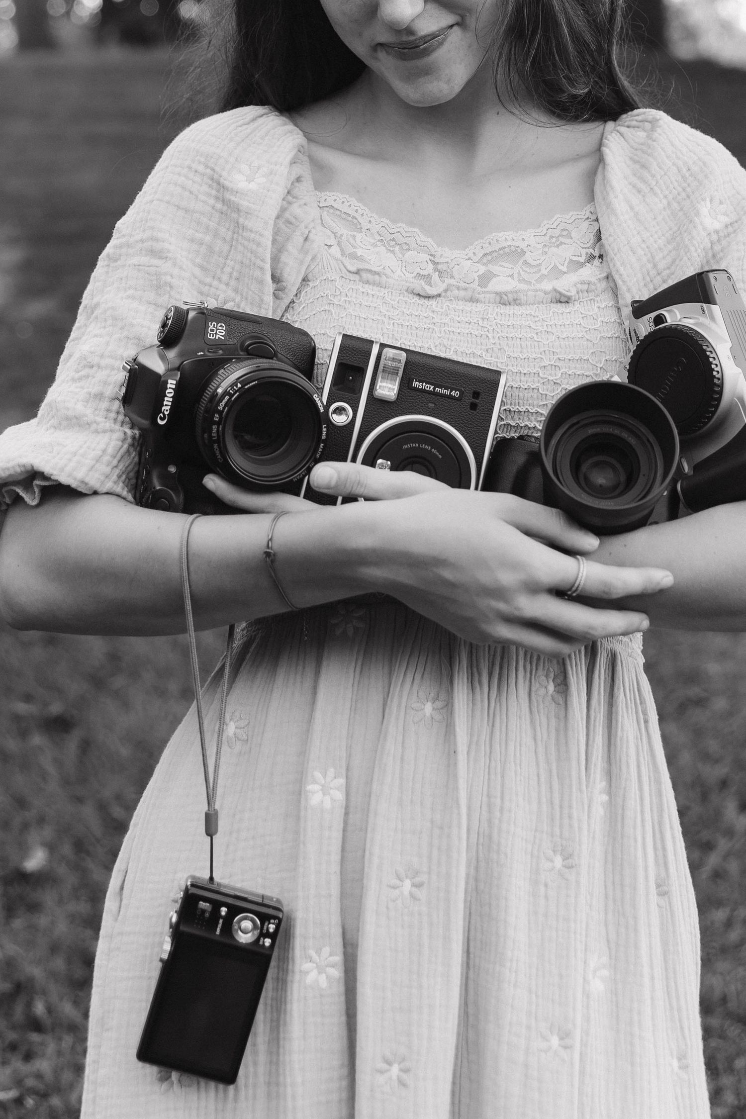 Woman in floral dress holding multiple cameras including Canon and Fujifilm Instax Mini in black and white photo.