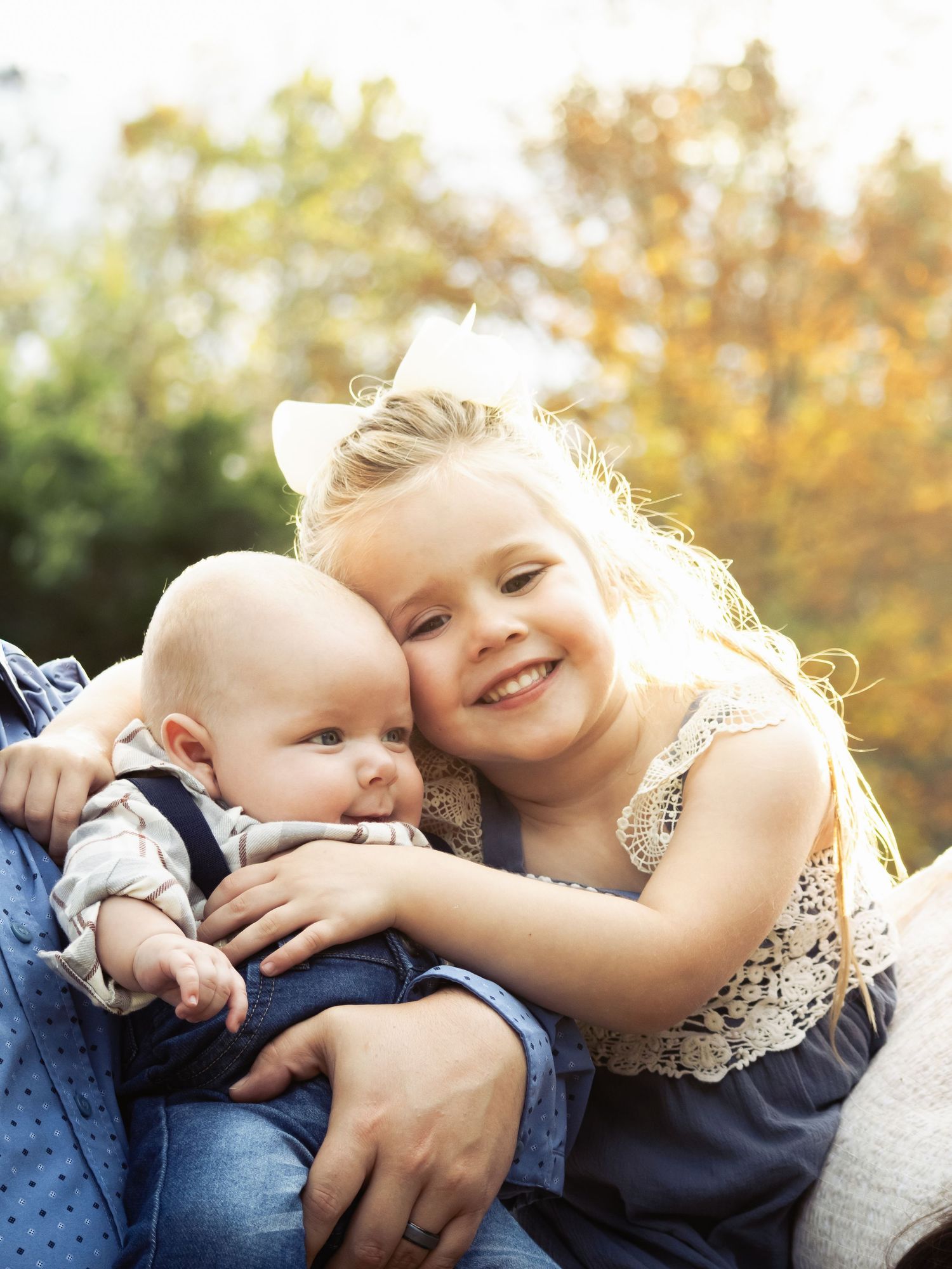 A smiling young girl with blonde hair hugs a baby outdoors with autumn trees in the background.