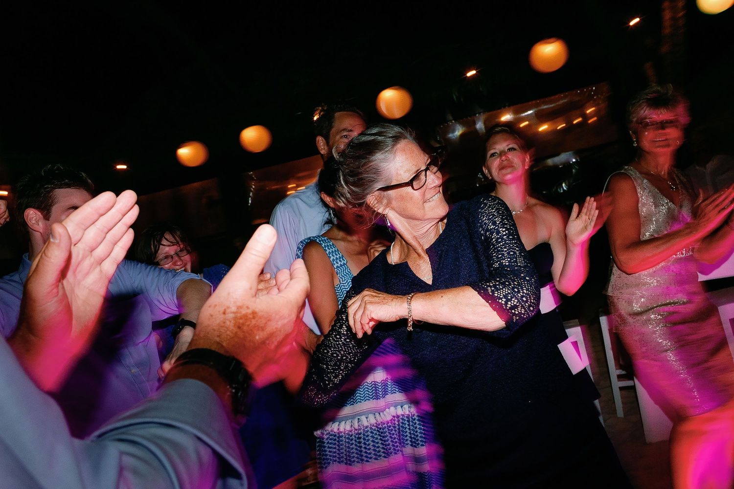 A guest in a navy lace dress dances among friends beneath colorful reception lights.