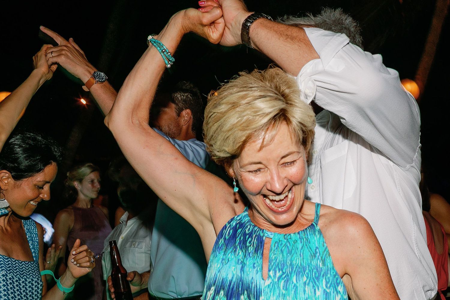 A guest in a blue patterned dress raises her arms and laughs on the crowded wedding dance floor.
