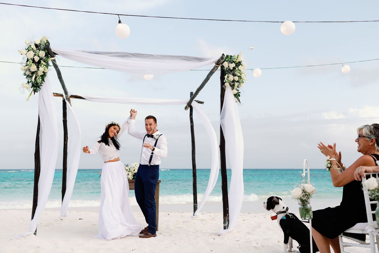 ewly married couple celebrating beneath a floral arch at the end of their Xpu-Ha beach wedding ceremony as their guests and dog look on.