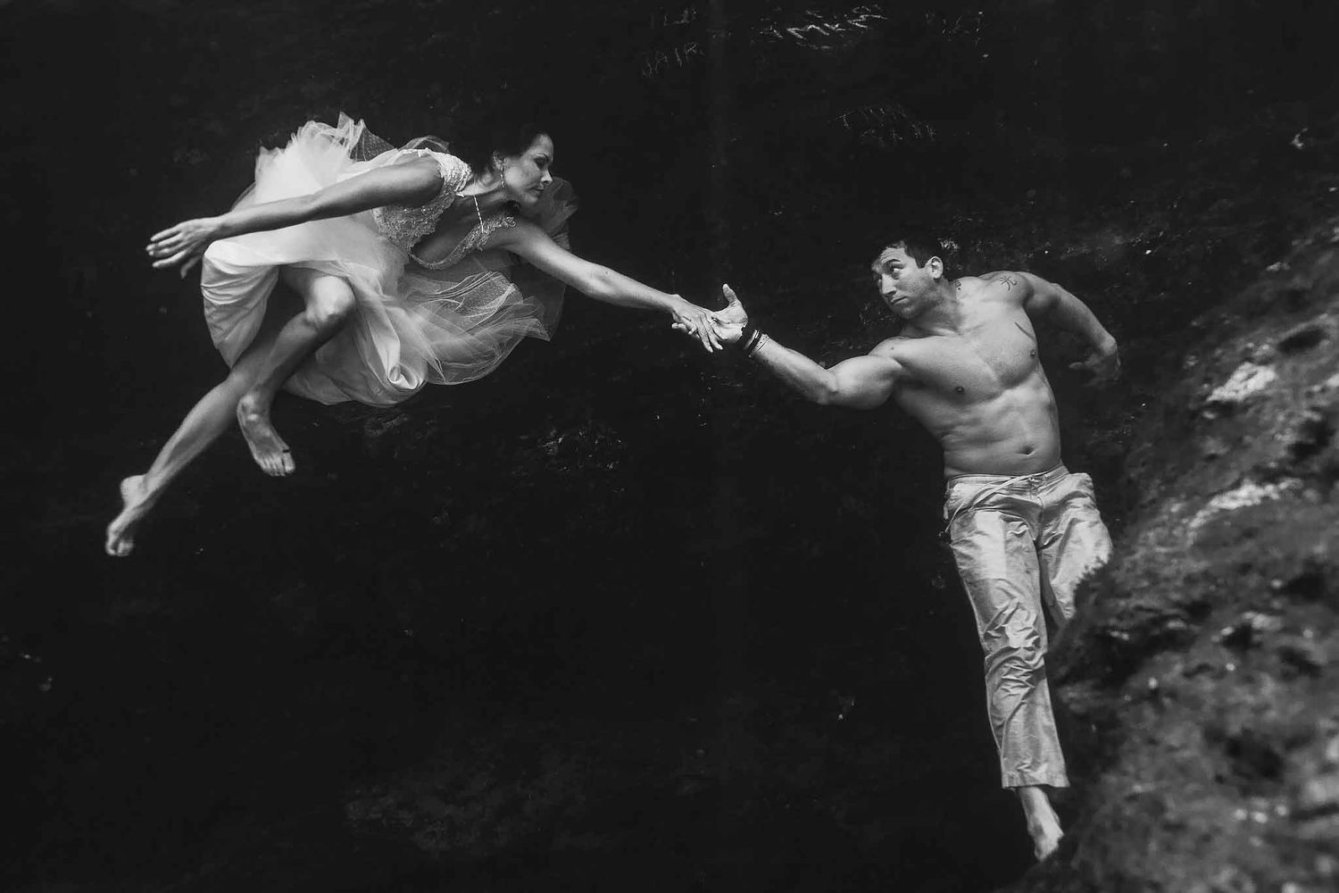 Fine art underwater wedding portrait of a bride and groom reaching for each other inside a Riviera Maya cenote in black and white.