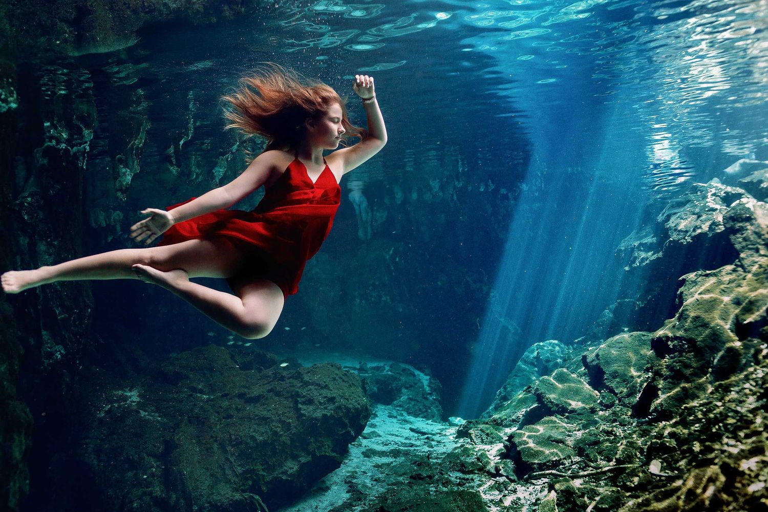 Underwater senior portrait photography session in a Tulum cenote featuring a young woman in a flowing red dress surrounded by crystal-clear
