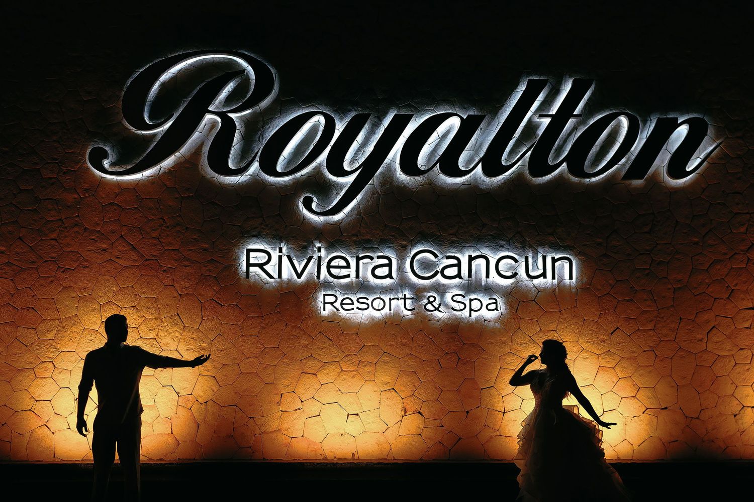 Silhouettes of the bride and groom posing beneath the illuminated Royalton Riviera Cancun Resort & Spa sign during a creative nighttime port