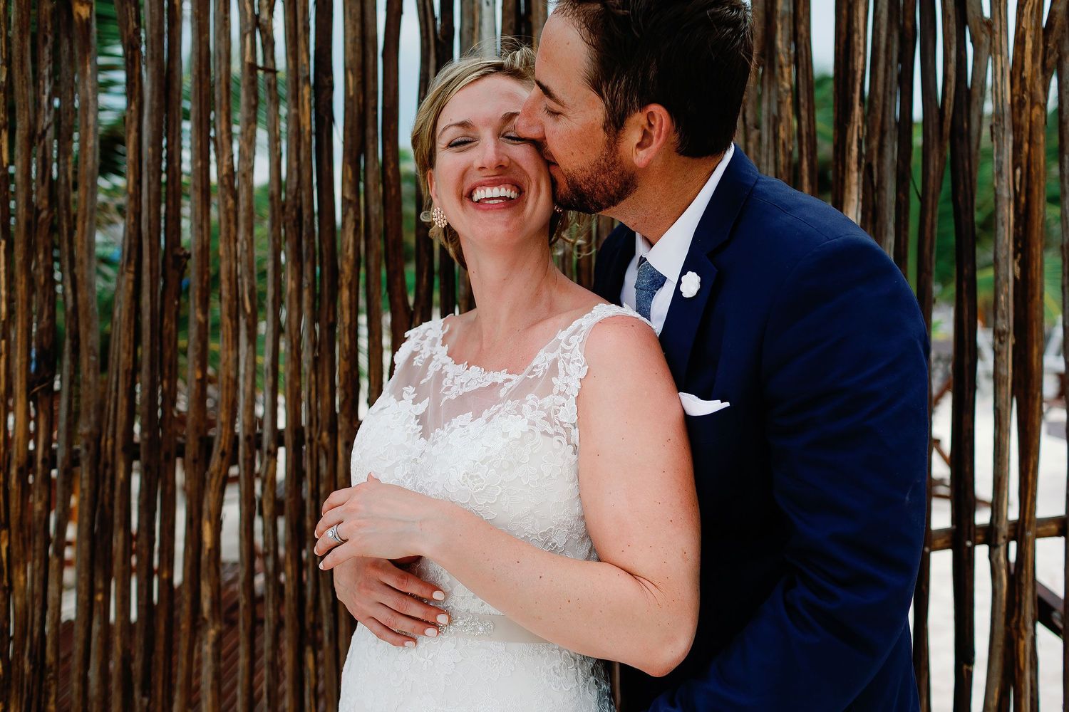 Matt kisses a laughing Ashley beside a wall of natural wooden branches in Tulum.