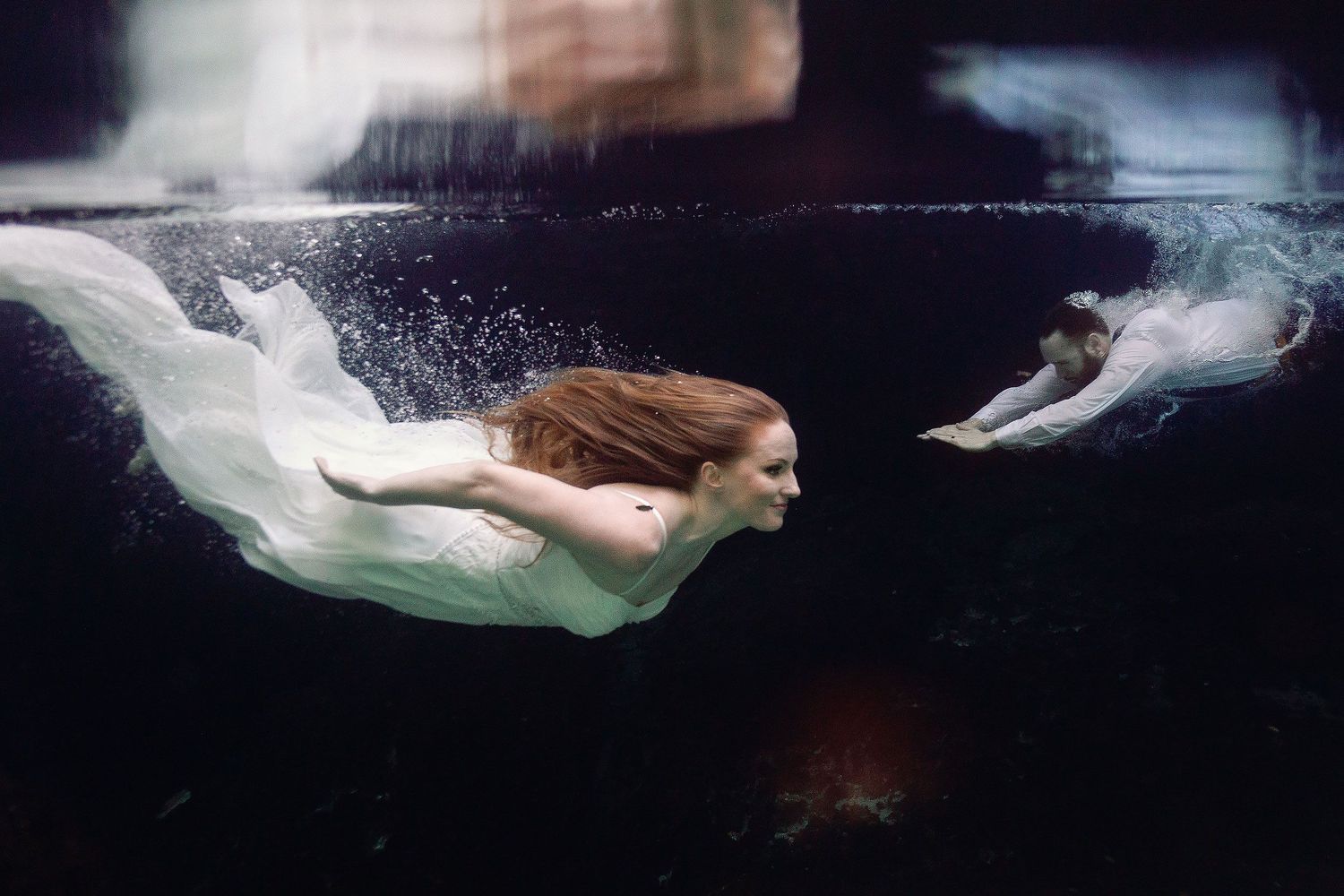 Nicole swimming through the clear water as Thomas follows behind during their underwater wedding portrait session at Cenote Azul near Playa