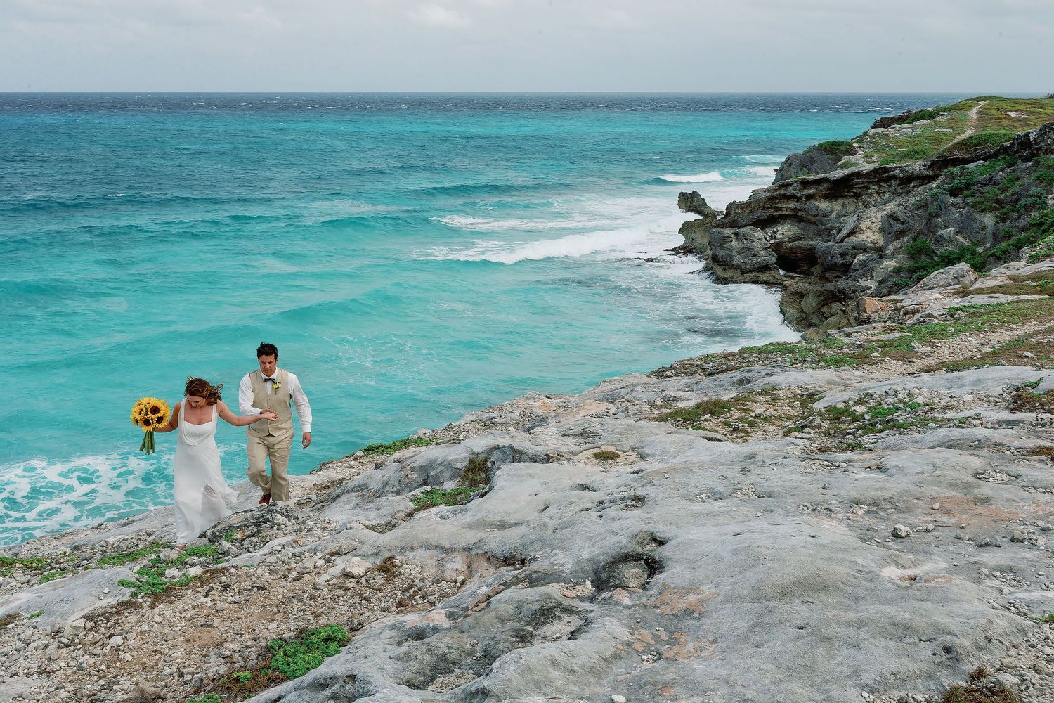 Li and Carlos climb a rocky Isla Mujeres shoreline hand in hand above turquoise Caribbean water.