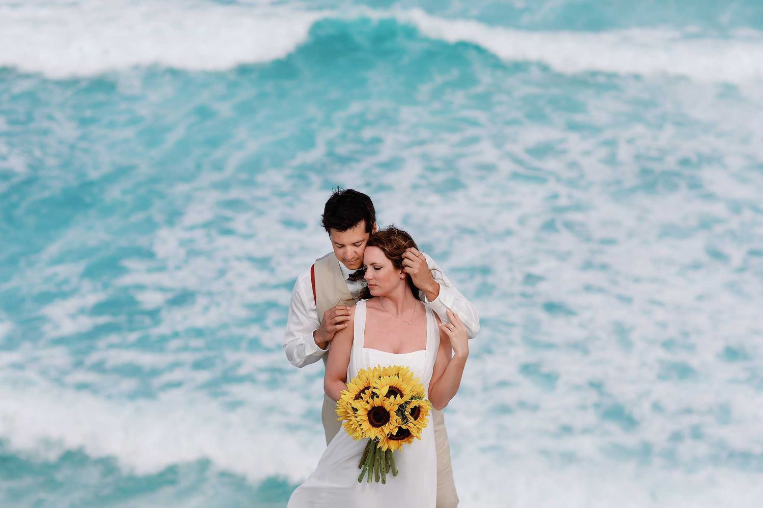 Romantic beach wedding portrait in Isla Mujeres with couple embracing by turquoise Caribbean waves holding sunflower bouquet