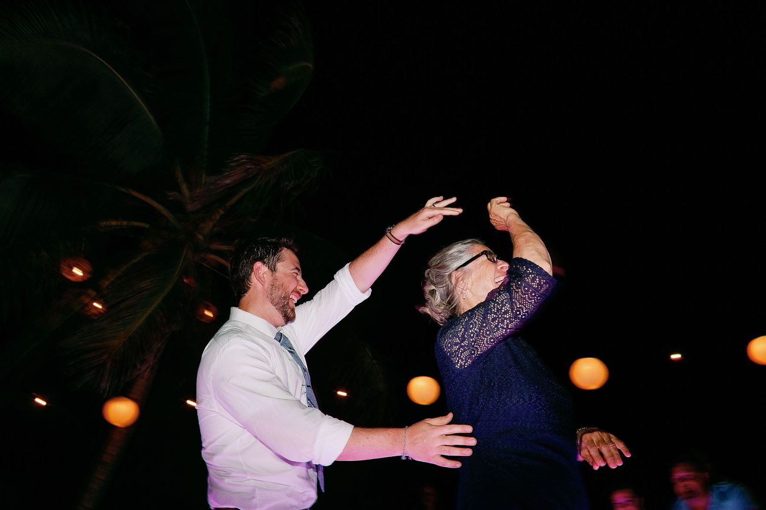 Matt laughs and dances with a guest beneath palm trees and glowing lanterns at the reception.