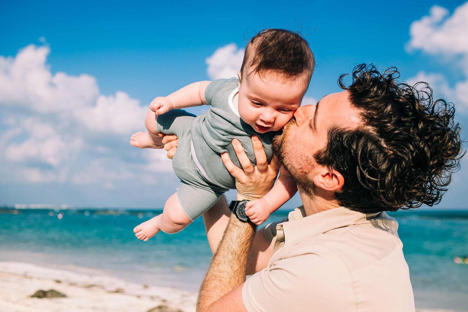 Father holding and kissing baby during a tropical family photography session at Fairmont Mayakoba beach.
