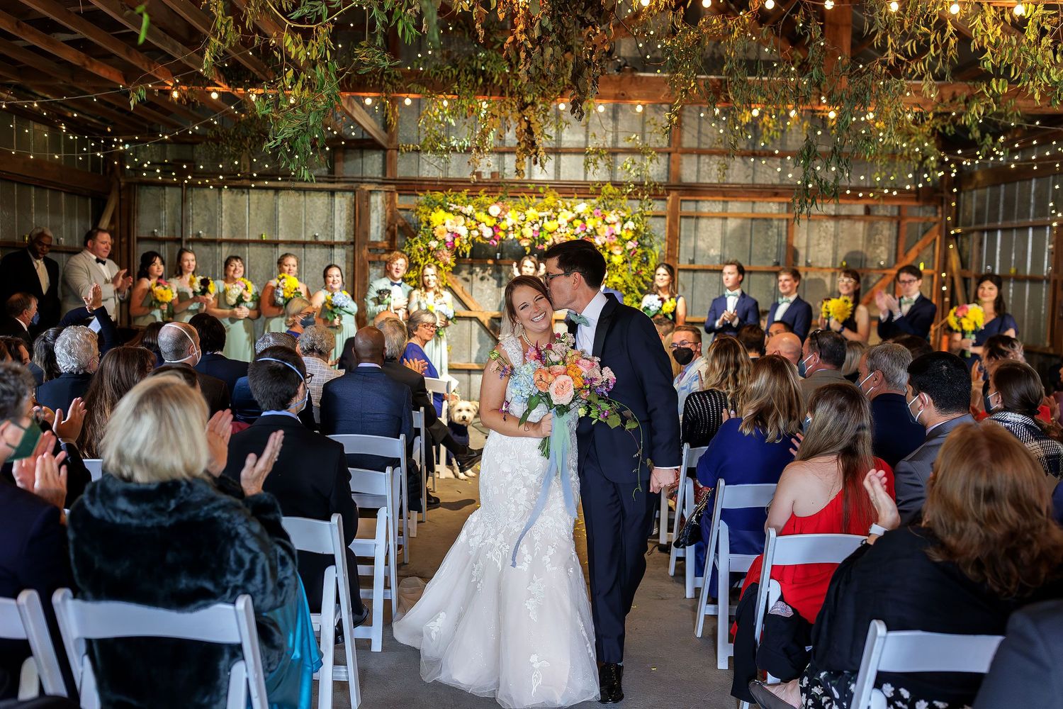 Groom kissing the smiling bride’s temple as guests applaud during their ceremony exit in a barn decorated with greenery and string lights.