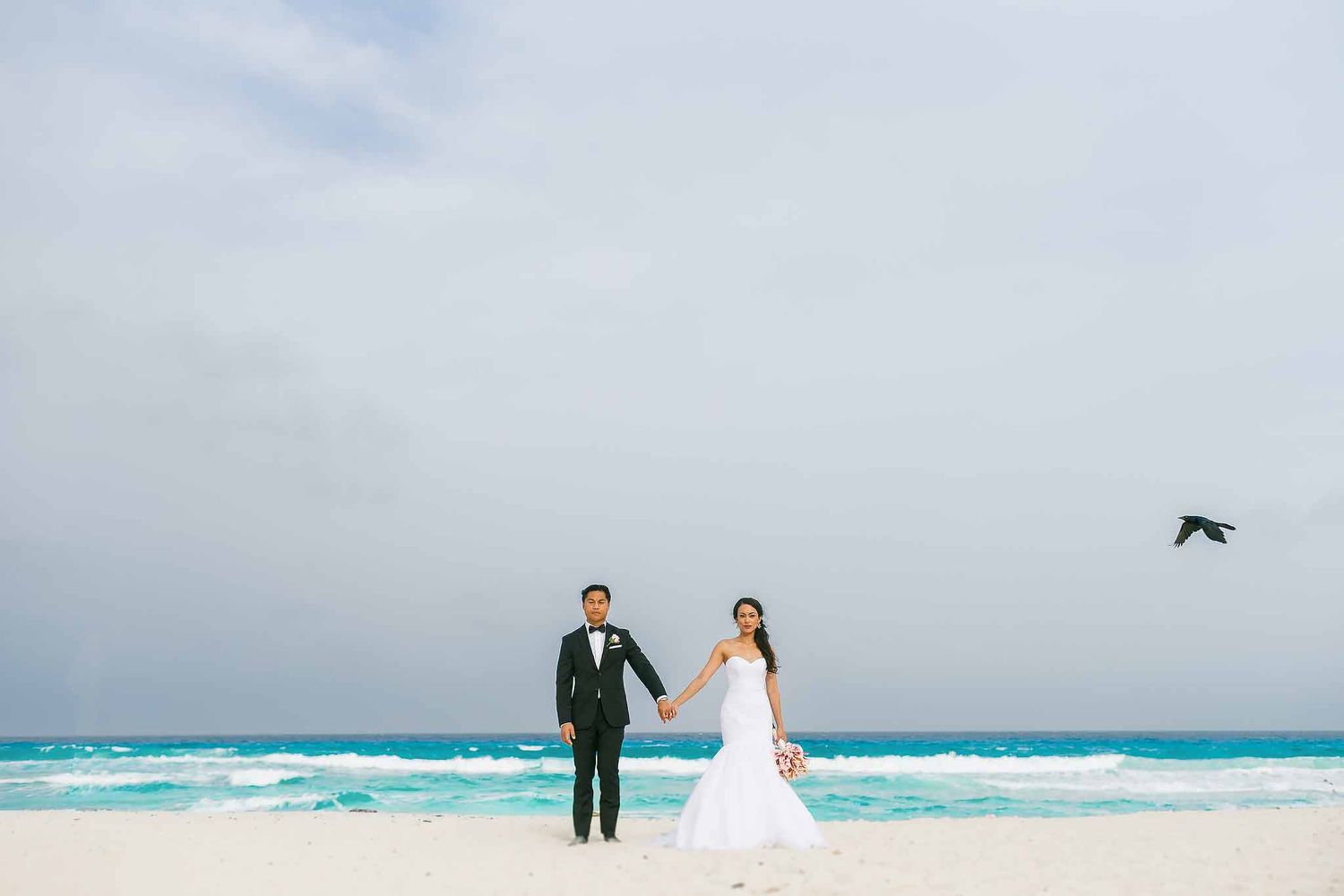 Bride and groom posing on a white sand beach in Cancun with turquoise Caribbean waves and dramatic sky during destination wedding portraits.