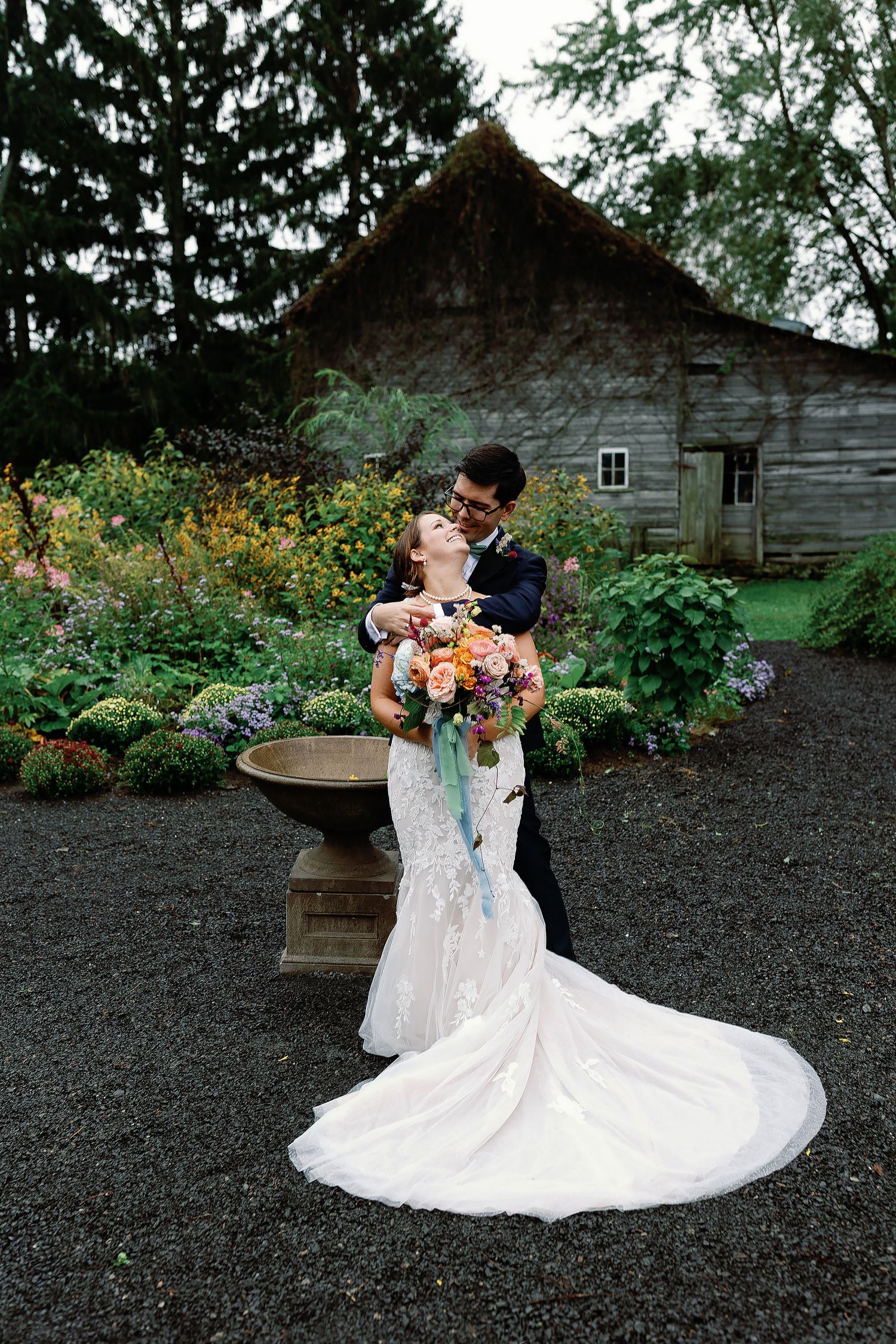 Groom embracing the bride from behind as she holds a colorful bouquet in a flower garden beside a weathered wooden building.