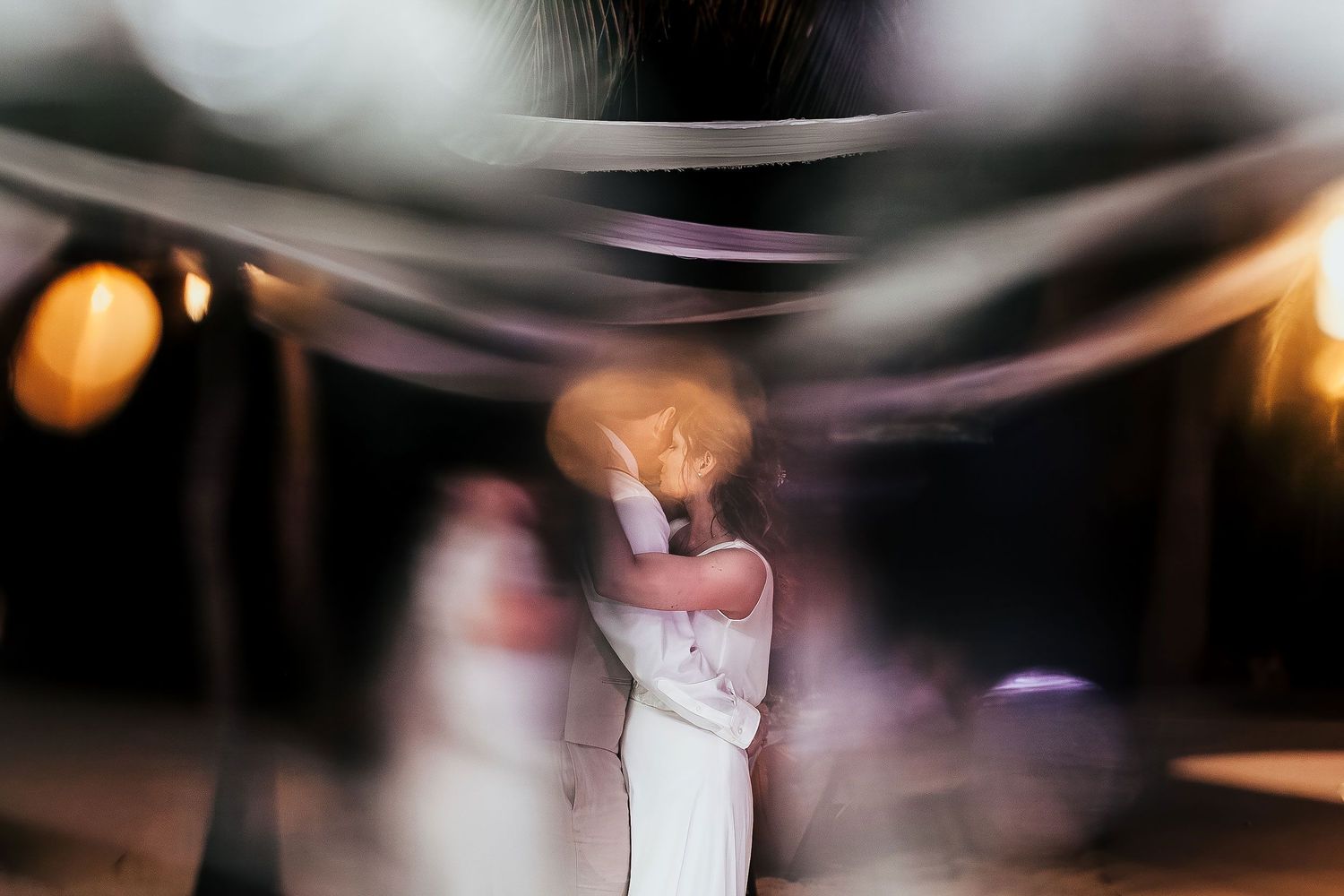 Creative nighttime portrait of Li and Carlos kissing beneath flowing fabric at their beach reception.