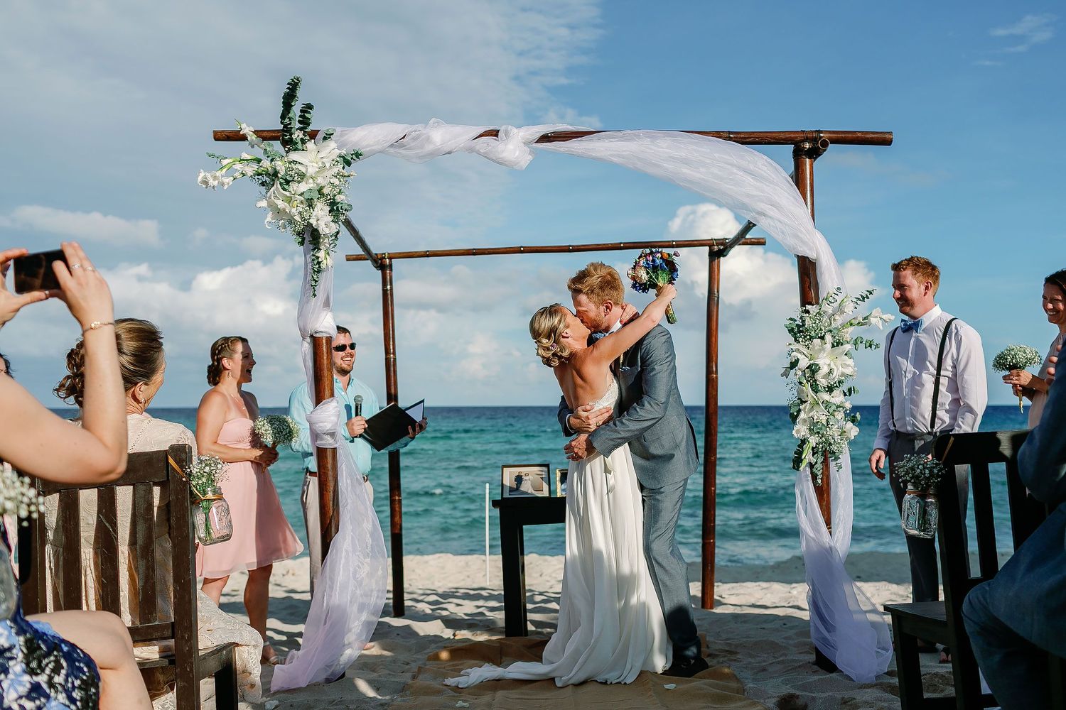 Cressida and Alan sharing their first kiss beneath a white floral arch during their private beach wedding ceremony in the Riviera Maya.