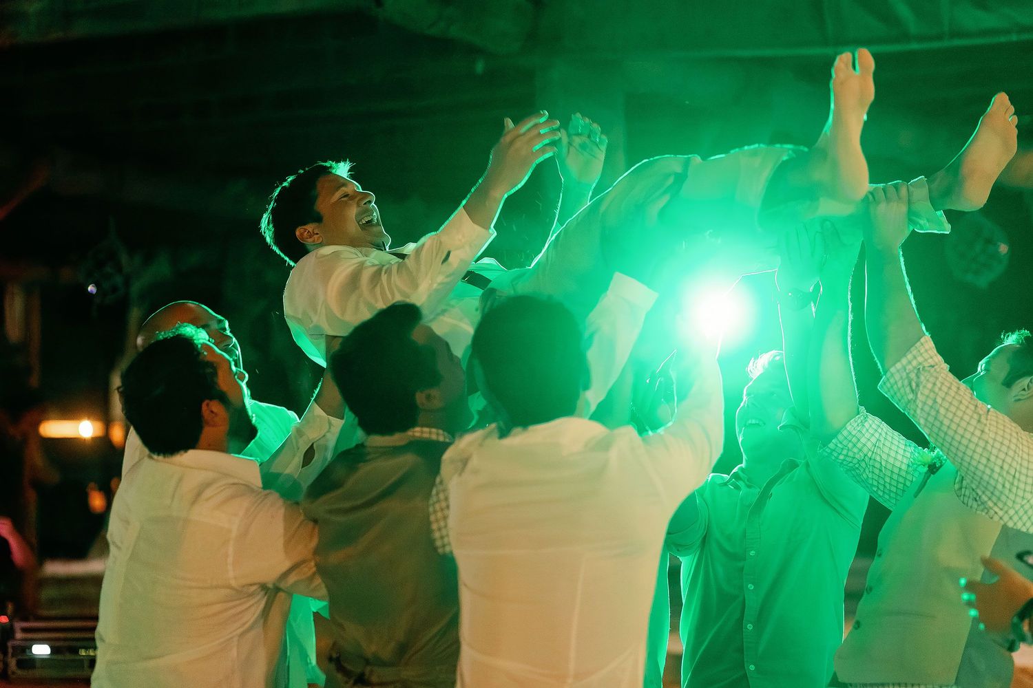 Guests lift Carlos above the dance floor beneath vivid green reception lighting.