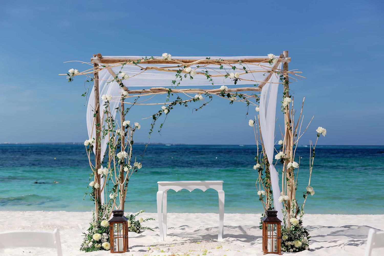 Elegant beach wedding arch setup at Riu Costa Mujeres with white flowers, wooden altar, turquoise Caribbean ocean, and white sand.