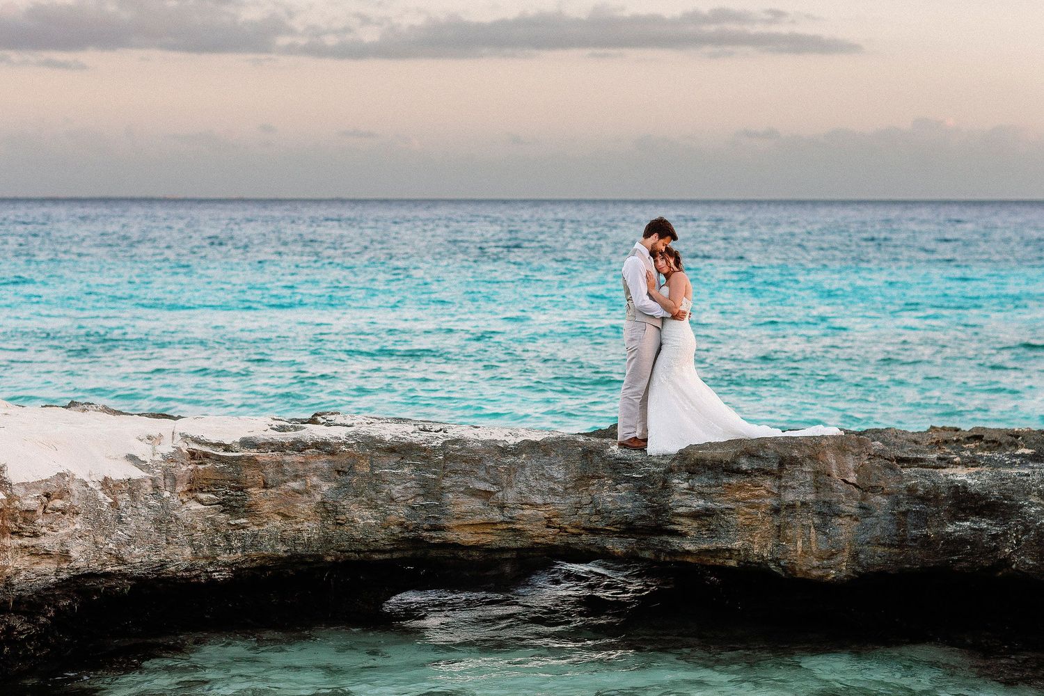Bride and groom embracing on a natural rock formation above the turquoise Caribbean Sea during their portrait session at Hotel Xcaret Mexico
