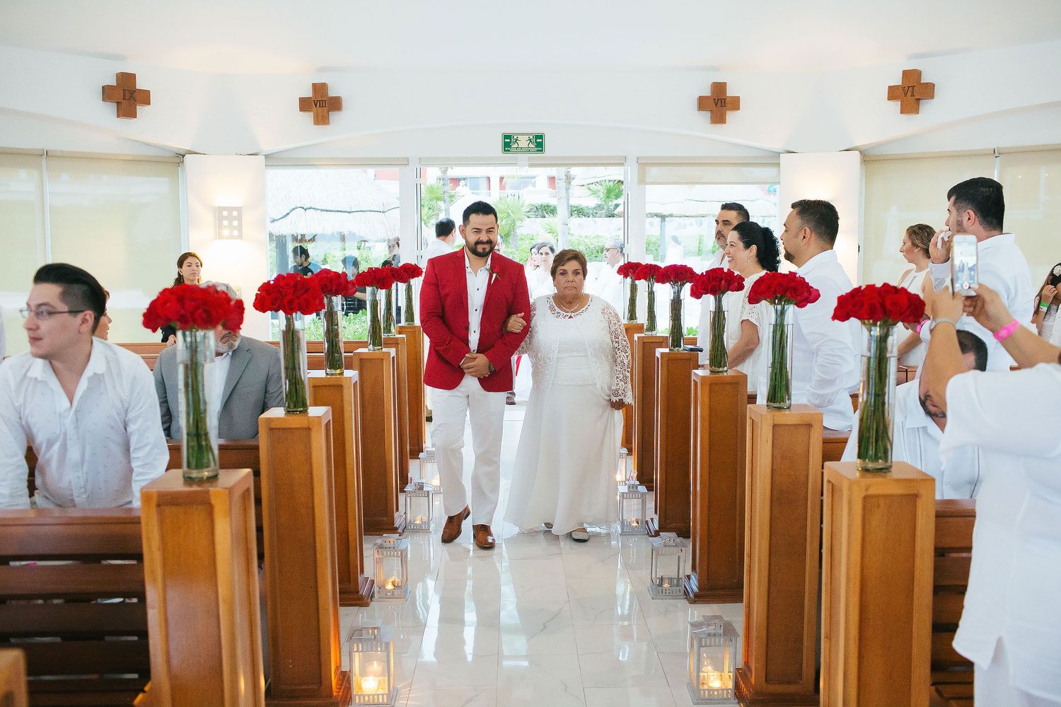 Wedding processional inside San Charbel Chapel at Hard Rock Riviera Maya featuring red floral aisle decorations and family members