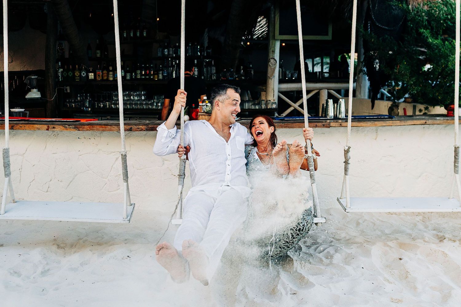 Sarah and Evish laugh together on beach swings after their Tulum elopement.