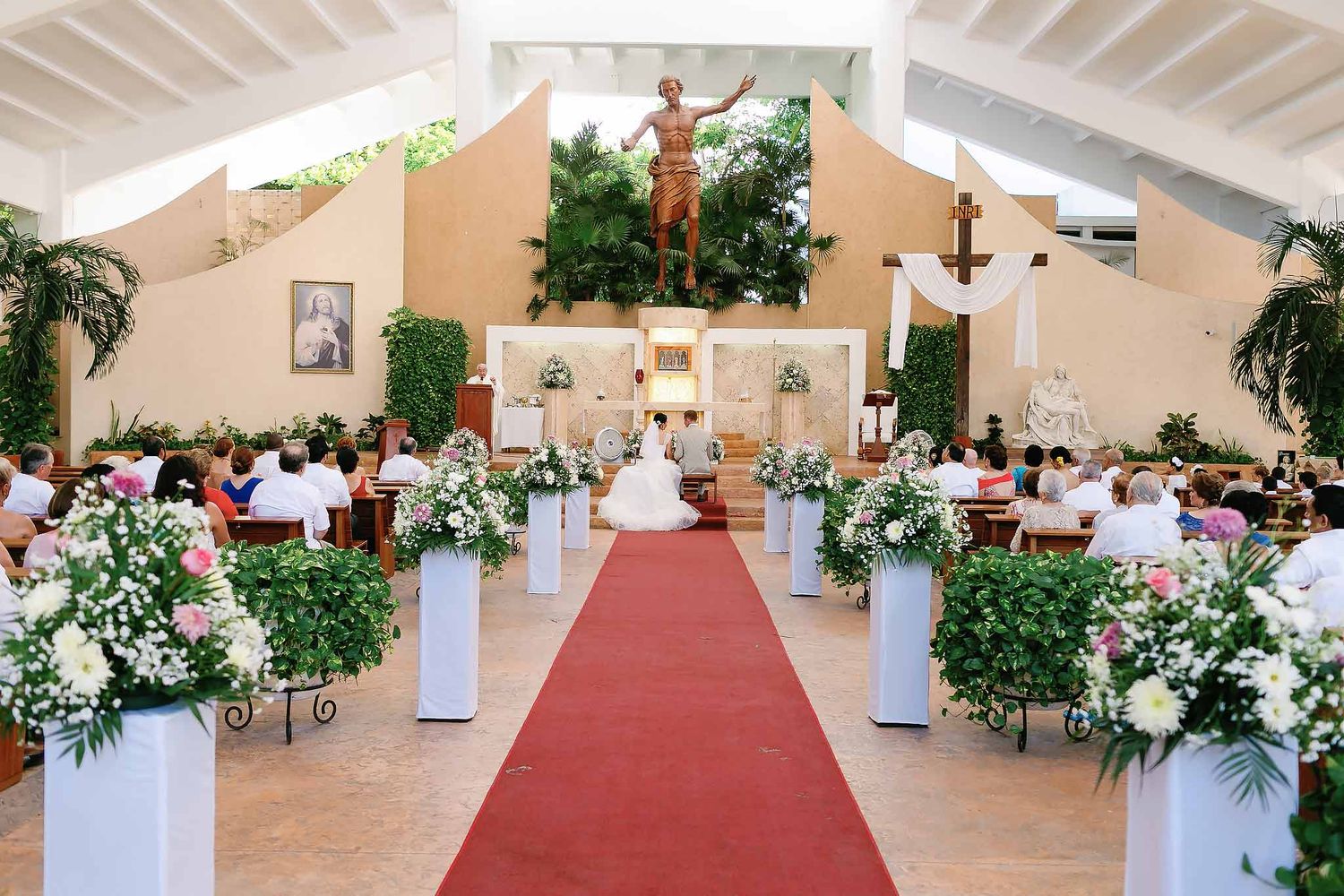 Wide interior view of Cristo Resucitado Chapel in Cancun Hotel Zone decorated for Catholic wedding with floral aisle and guests