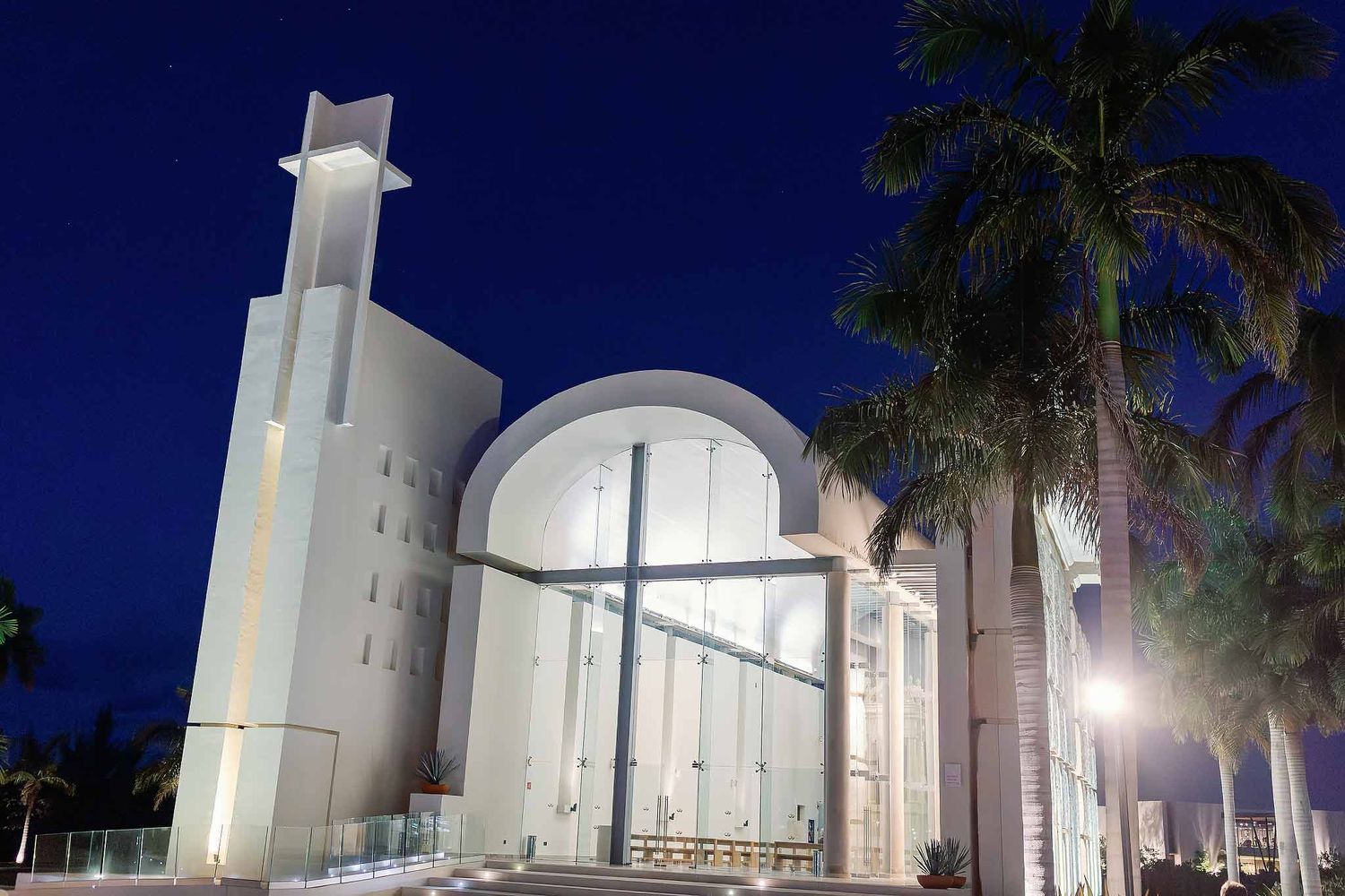 Exterior of Our Lady of Rocío Catholic chapel at Grand Palladium Costa Mujeres illuminated at night with palm trees