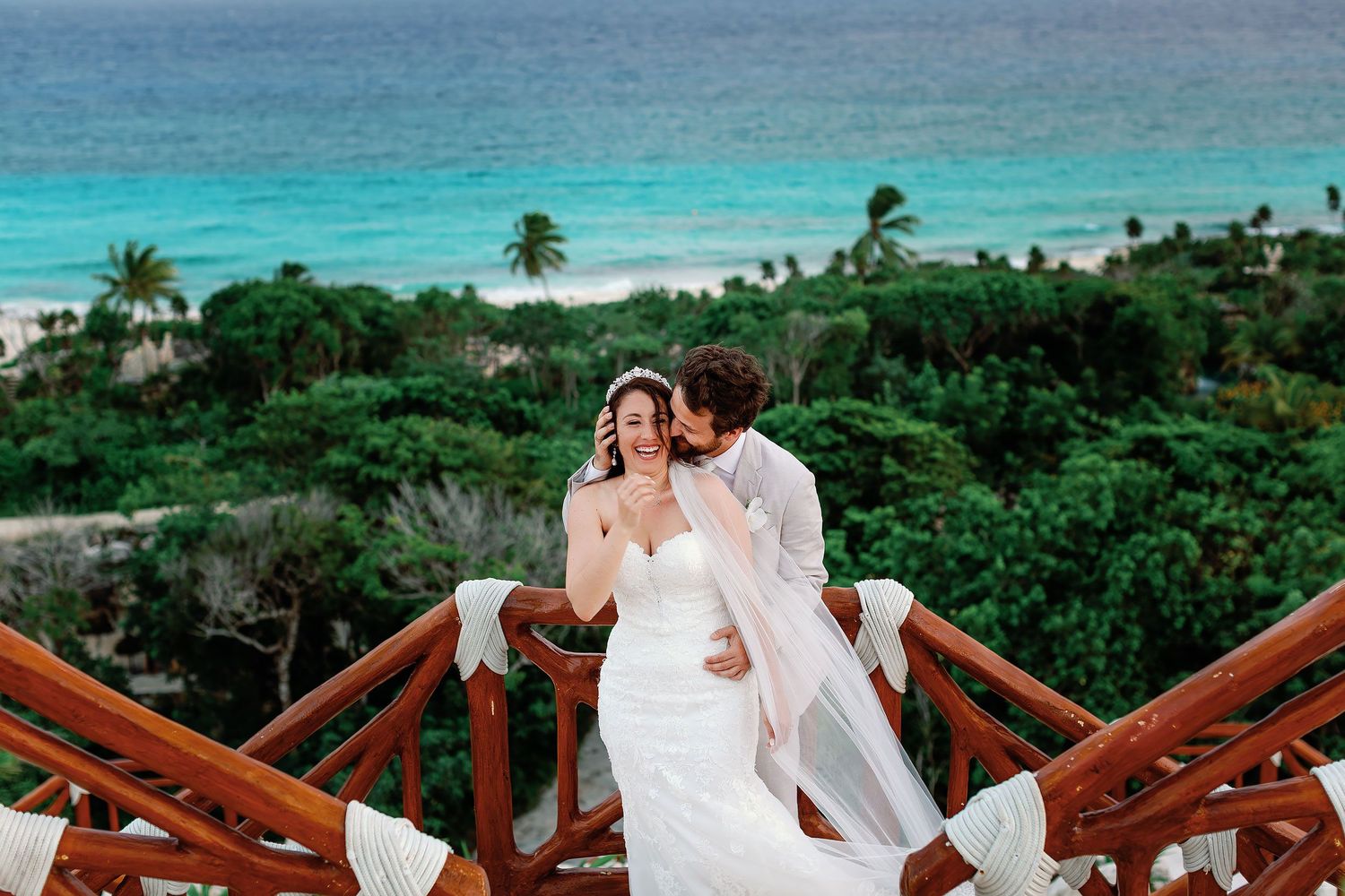Laughing bride and groom embracing on a wooden terrace overlooking the tropical jungle and Caribbean Sea at Hotel Xcaret Mexico.
