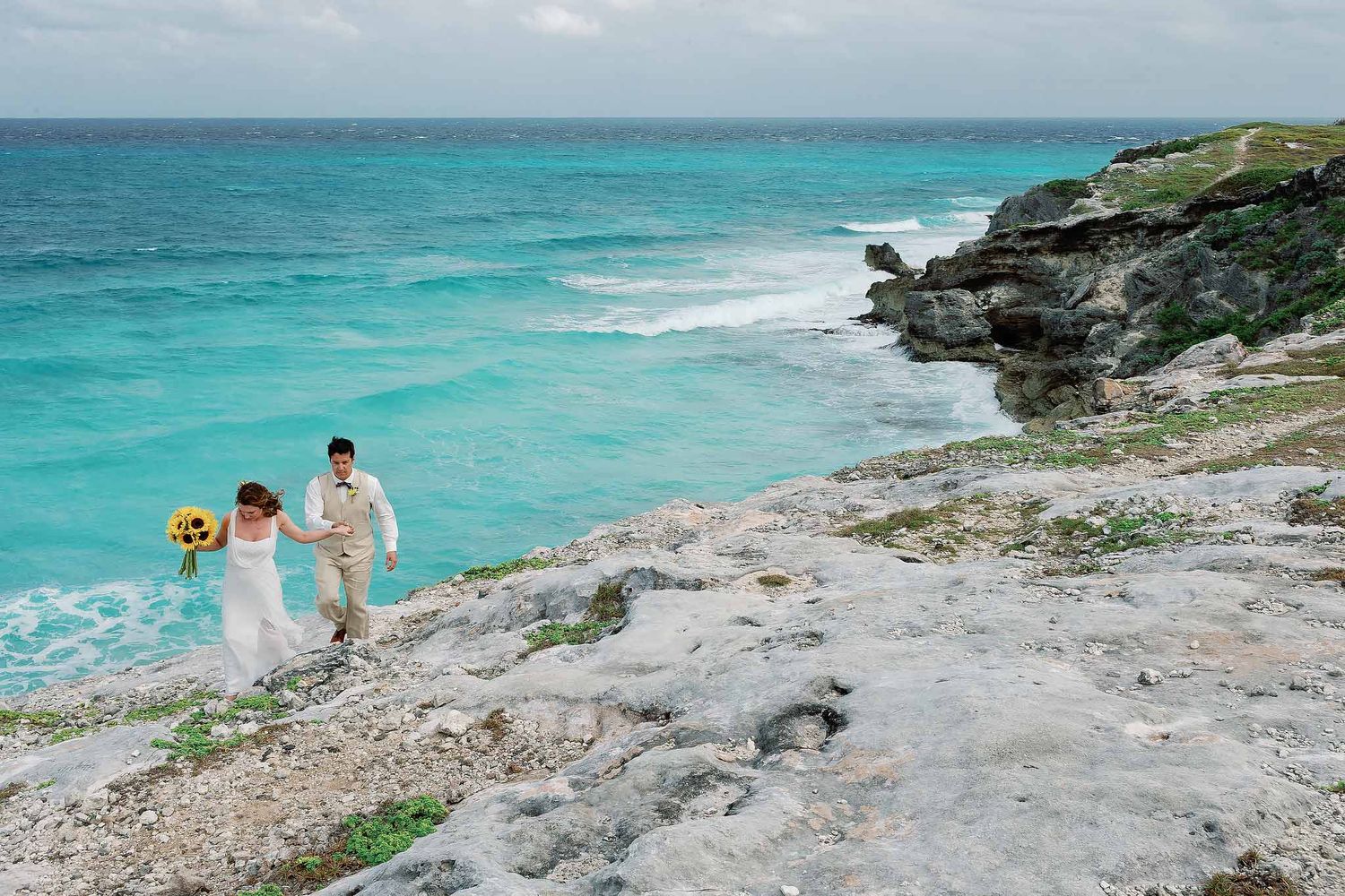 Bride and groom walking along Cancun cliffside coastline with turquoise Caribbean waters during a destination wedding photography session.