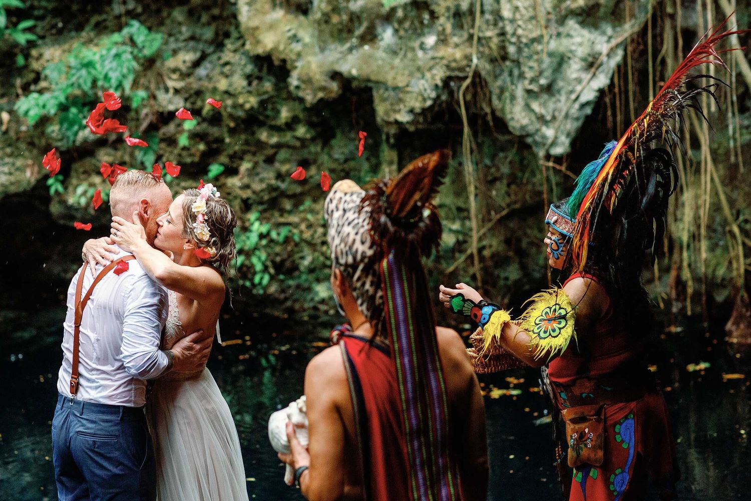 Jeff and Veronica kissing beneath falling red petals at the end of their Maya wedding ceremony at Cenote Hats’uts near Playa del Carmen.