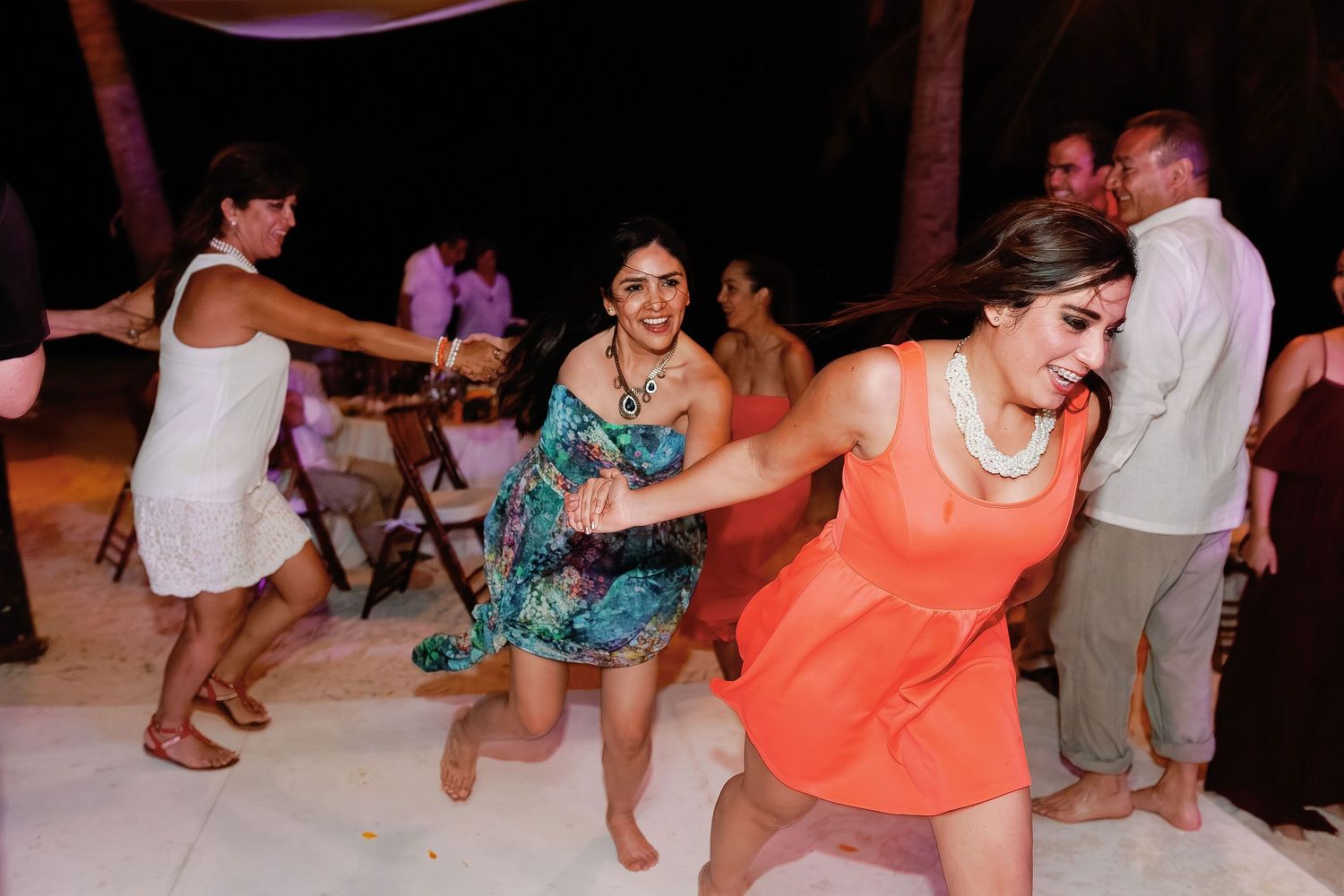 Women in colorful dresses hold hands and dance barefoot during the Playa Norte wedding reception.