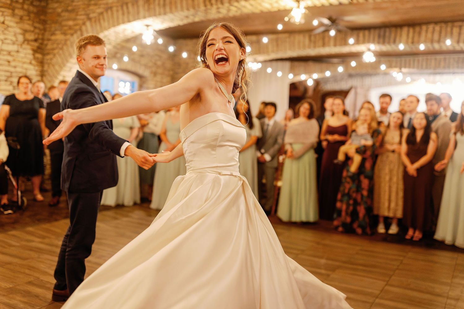 Kaylee laughing while dancing with Joseph as guests watch beneath string lights at Mayowood Stone Barn in Rochester, Minnesota.