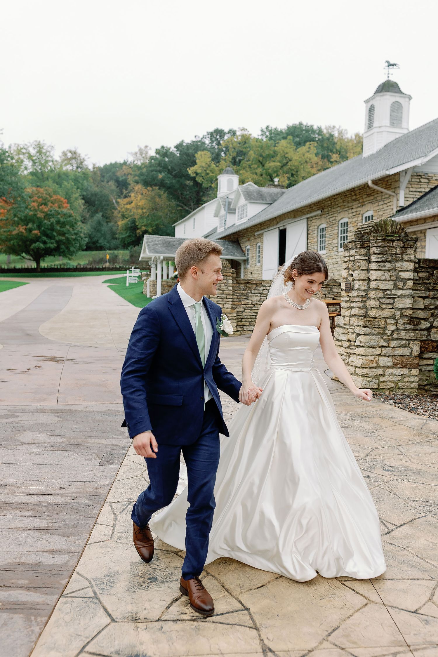 Kaylee and Joseph smiling and walking hand in hand outside Mayowood Stone Barn during their wedding in Rochester, Minnesota.