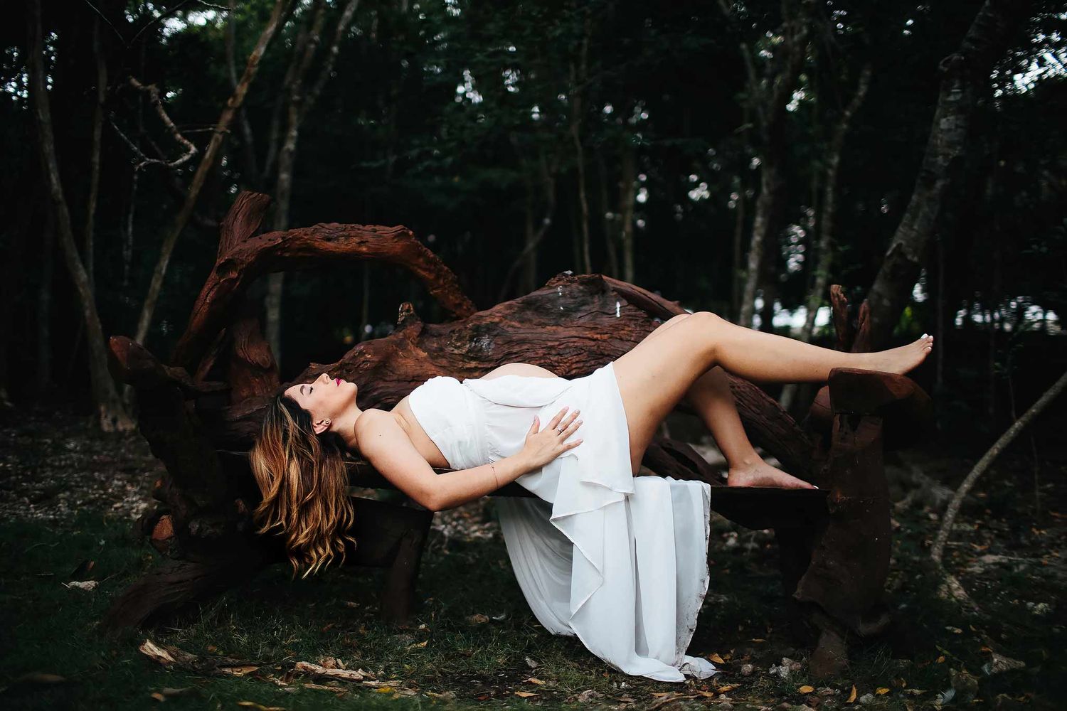 Pregnant woman in a flowing white dress reclining on a sculptural wooden bench during an artistic jungle maternity photography session in Tu