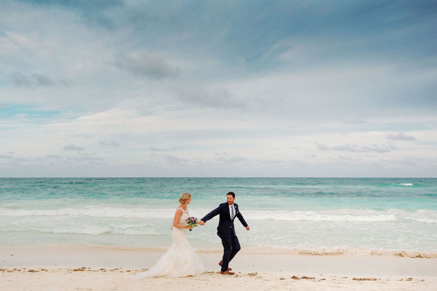 Ashley and Matt playfully run hand in hand beneath a dramatic sky on the Tulum beach.